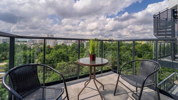 Un balcon avec deux fauteuils en osier, une petite table ronde ornée d’un pot de fleurs, et une vue sur la ville sous un ciel partiellement nuageux.