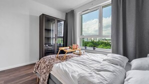 Une chambre avec un grand lit, une table de chevet en bois sur laquelle se trouvent une tasse et un livre, une armoire et une fenêtre donnant sur un paysage verdoyant.