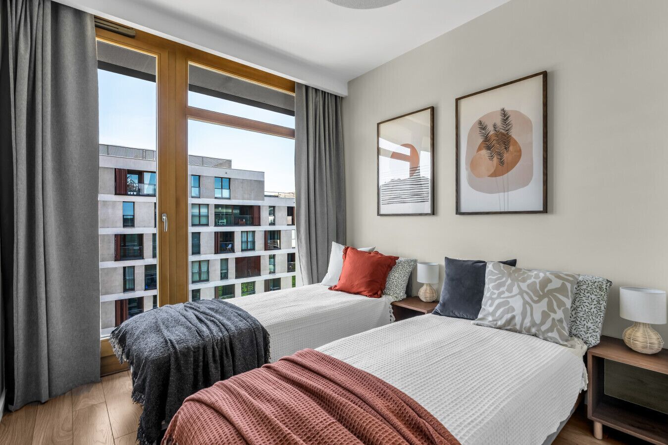 A hotel room with two beds, a large window with curtains, a view of buildings, and two framed artworks on the wall.