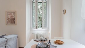 A dining area with a table set for two, a window with a view of trees, a framed menu on the wall, and a clock on the wall. - Nonna Tere Apartment (Carate Urio)