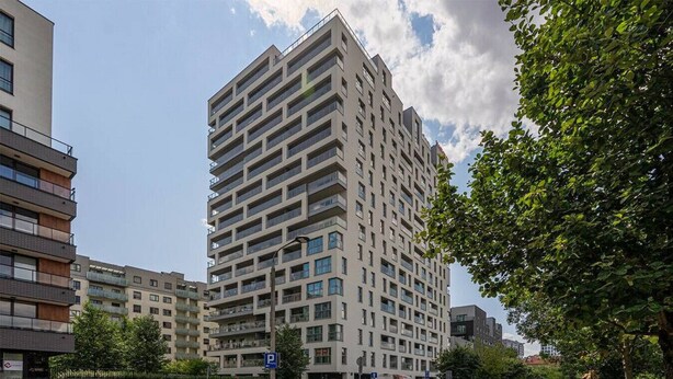 Un immeuble moderne de grande hauteur, doté de nombreux balcons, d’un espace de stationnement et surplombant un ciel dégagé.