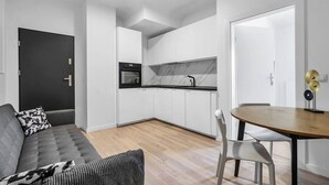 Une cuisine moderne avec des meubles blancs, un four noir et un pare-feu en marbre. On y trouve une table à manger avec deux chaises et un canapé à motifs noirs et blancs.
