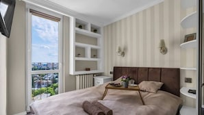 A bedroom with a large bed, a view through a window, built-in shelves, and a bedside table. - Warsaw Praga View - 17th Floor with Balcony - by Rentujemy (Warszawa)