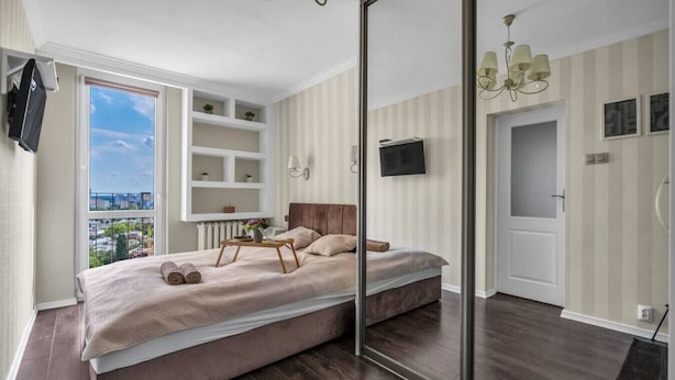 A bedroom with a large bed, a TV mounted on the wall, a window with a city view, and a mirror. - Warsaw Praga View - 17th Floor with Balcony - by Rentujemy (Warszawa)