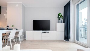 En moderne stue med et fladskærms-tv, hvide møbler og et stort vindue med gardiner.