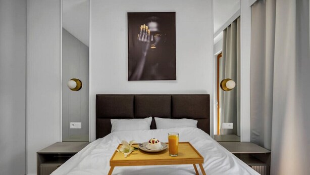Una habitación de hotel moderna con una cama grande, una mesita de noche con una bandeja de desayuno y un ventanal con cortinas.