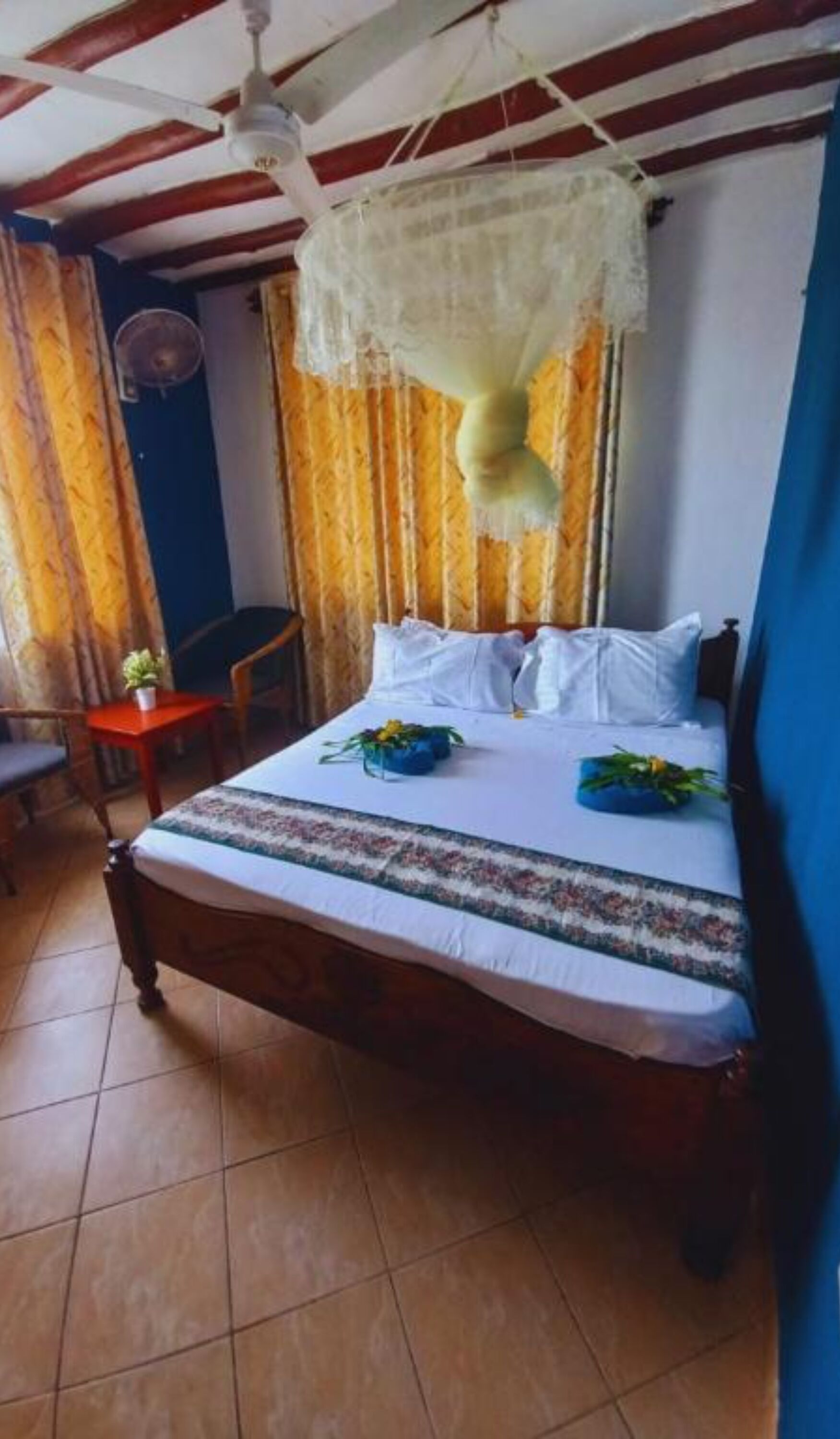 Un dormitorio con cama, una mesita con planta, una silla y un ventilador de techo.