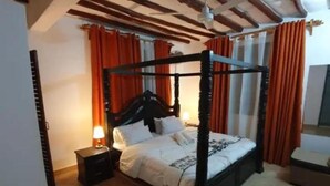 A four-poster bed with a wooden frame, a bedside table with a lamp, and orange curtains. - Casa Villa Diani (Diani Beach)