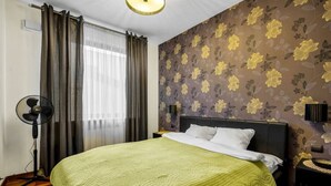 A hotel room with a bed, a standing fan, a bedside table, a nightstand, a lamp, and a floral wallpaper.