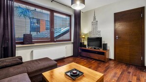 A modern living room with a sofa, a wooden coffee table, a TV on a wooden stand, and a large window with blinds.