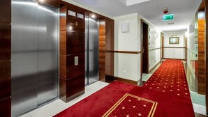 A hotel hallway with elevators, a red carpet, and a green exit sign.