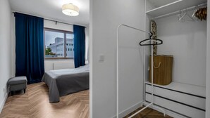 A bedroom with a bed, a small bench, a window with curtains, a radiator, and a wardrobe with a hanging rack. - Business District Apartment - Parking, Balcony by Rentujemy (Warszawa)