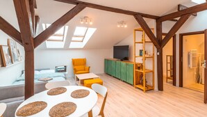 A cozy loft-style room with exposed wooden beams, a dining area with a white table and woven placemats, a bright yellow armchair, and a green cabinet with a TV. - Kraków Comfort Loft Apartments by Rentujemy (Kraków)