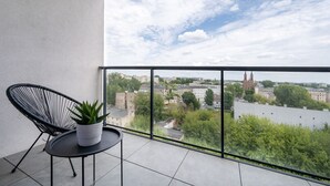 Un balcon avec une chaise noire et un pot de fleurs, donnant sur un paysage urbain avec des immeubles et une rivière.