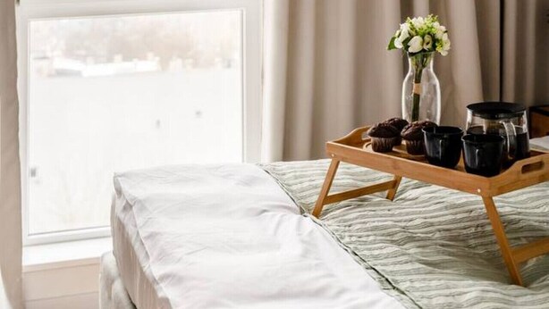 Une chambre avec un lit, une fenêtre donnant sur un paysage urbain, un plateau avec des tasses à café et des muffins, et un vase rempli de fleurs.