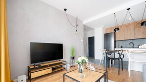 Un salon moderne doté d’un téléviseur à écran plat, de parquet, d’une table basse et d’un coin repas avec des chaises blanches, et une cuisine en arrière-plan.