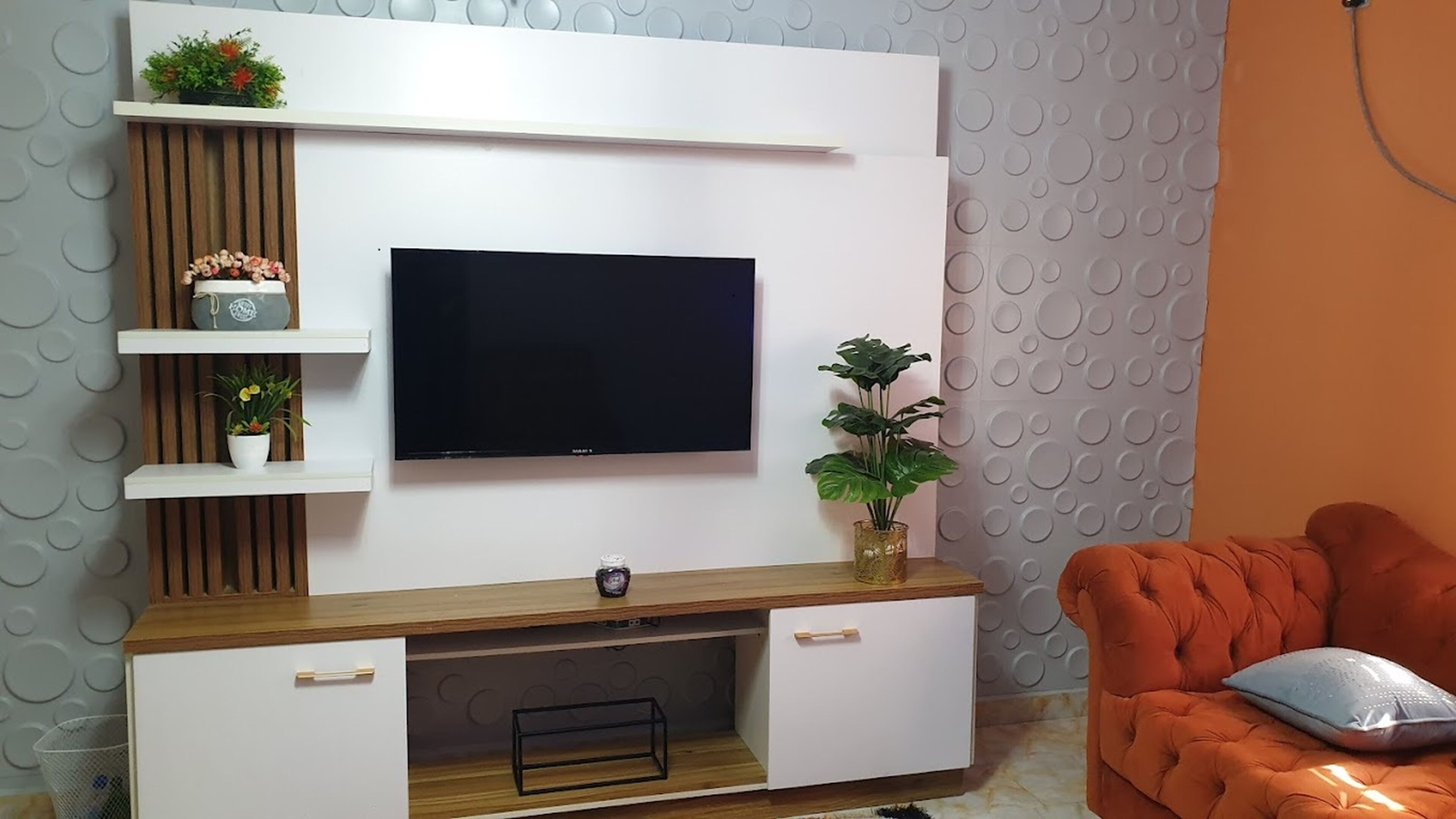 A modern living room with a flat-screen TV mounted on a white wall, wooden shelves with decorative items, a wooden cabinet with a TV stand, and a plush orange sofa.
