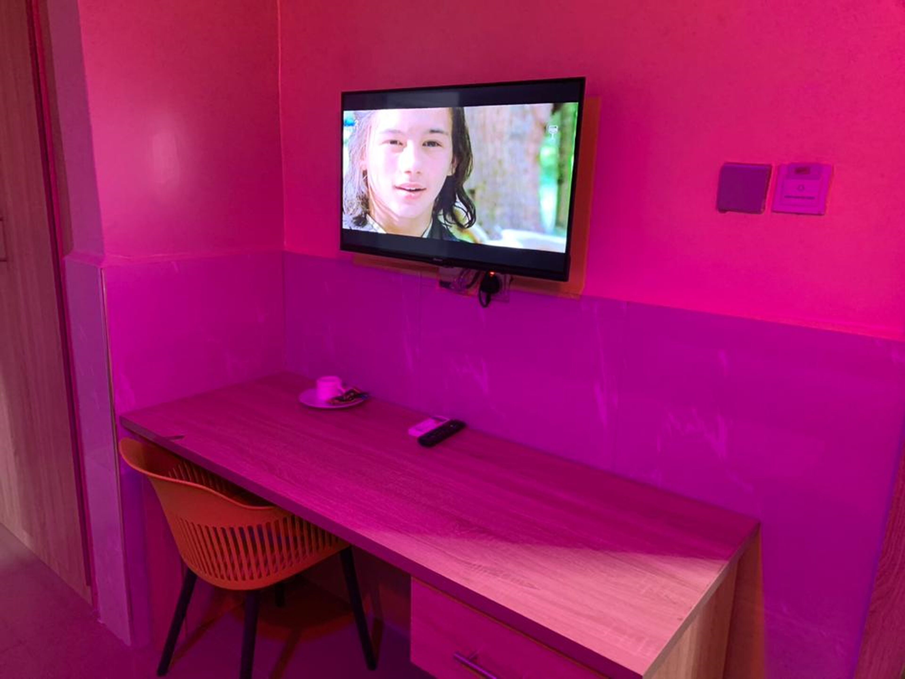 Una habitación con pared rosa, televisor montado, escritorio de madera, una silla y una mesita con una taza y un control remoto.