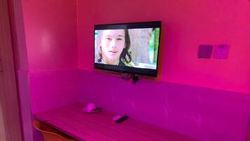 Una habitación con pared rosa, televisor montado, escritorio de madera, una silla y una mesita con una taza y un control remoto.