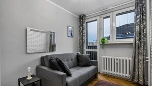 Een moderne woonkamer met een grijze bank, een klein bijzettafeltje, een radiator en een raam met gordijnen.