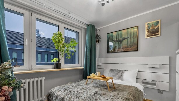 Een moderne slaapkamer met een groot raam, een zacht vloerkleed, een nachtkastje met dienblad en een schilderij aan de muur.