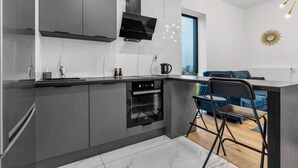 A modern kitchen with grey cabinets, a built-in oven, and a black range hood. There is a blue sofa and a folding chair in the adjacent living area. - Bliska Wola - City Skyline View, 10th Floor, Balcony - by Rentujemy (Warszawa)