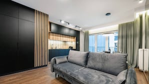Una sala moderna con un sofá gris, piso de madera, un armario negro y una zona de cocina con mostrador de madera.