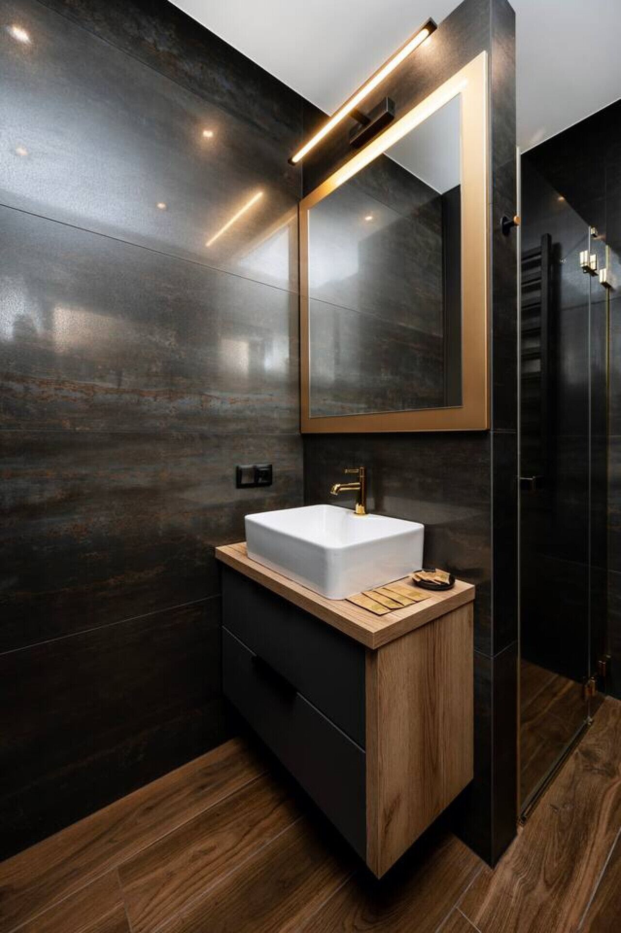 Un baño moderno con piso de madera oscura, un lavamanos blanco y un espejo grande con marco dorado.