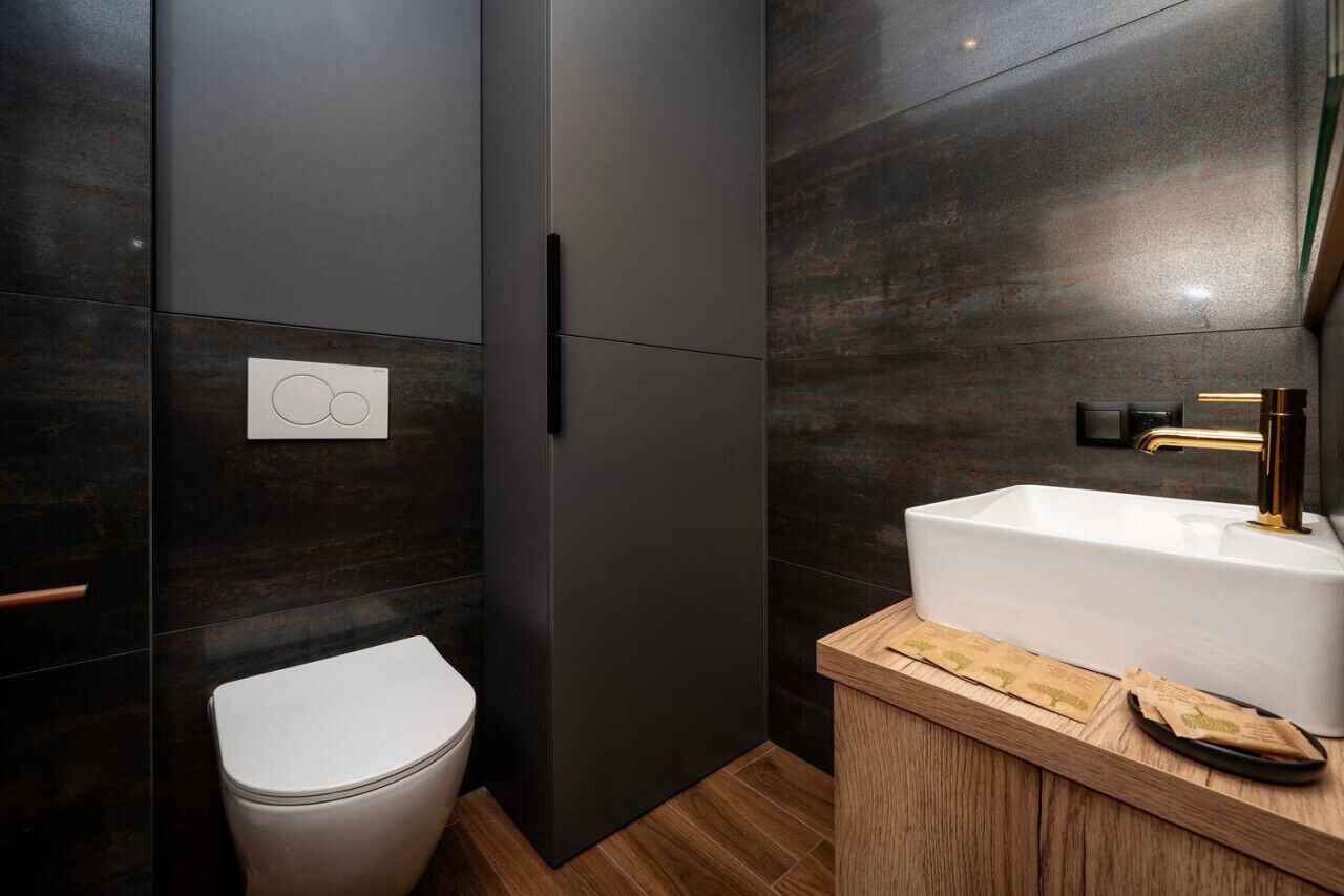 A modern bathroom with dark tiled walls, a white toilet, a wooden vanity with a sink, and a gold faucet.