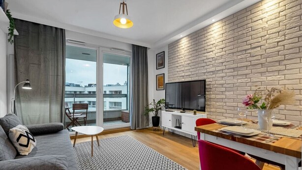A modern living room with a grey sofa, a dining table set for two, a TV, and a brick wall. - Warsaw Wola - Parking & Balcony - by Rentujemy (Warszawa)