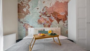 A bedroom with a world map mural on the wall, a bed with a white comforter, a wooden bedside table with a plate of food and a glass of orange juice, and a white radiator.