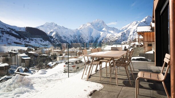 A snow-covered mountain landscape with a view of a town and a table with chairs on a balcony.
