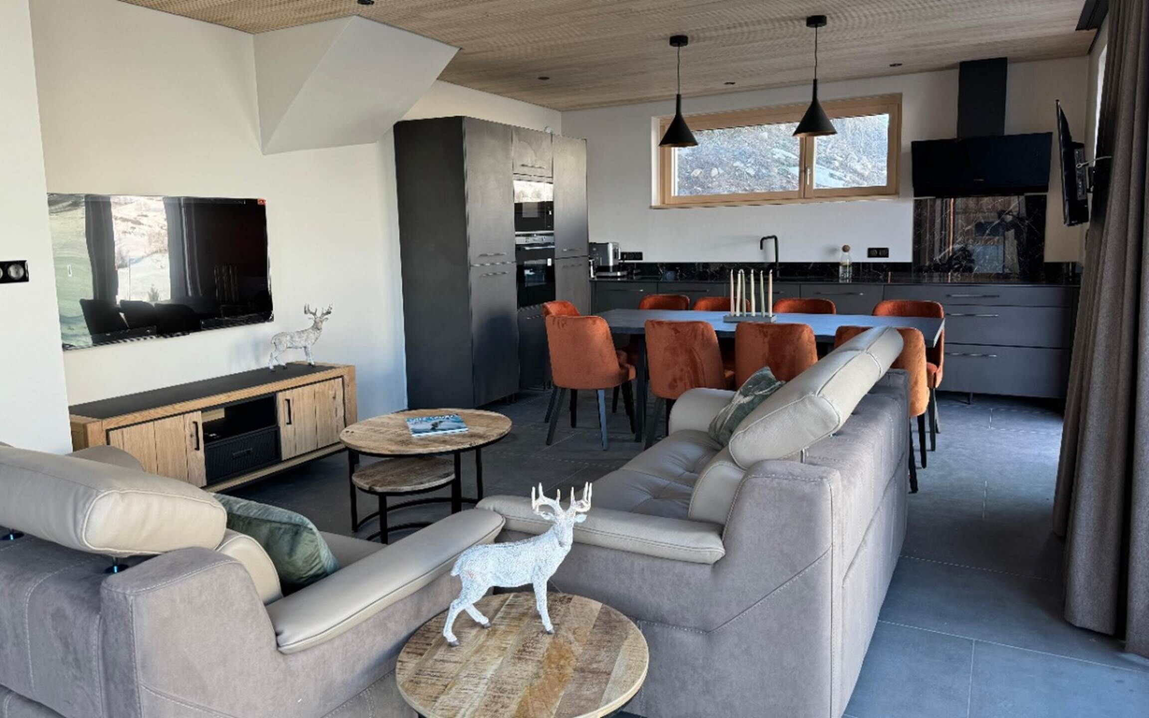 Una sala de estar moderna con un sofá gris, una mesa de centro y un televisor. Se observa una zona de cocina con una mesa de comedor y sillas.