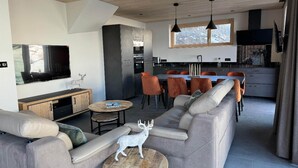 A modern living room with a grey sofa, a coffee table, and a TV. There's a kitchen area with a dining table and chairs.