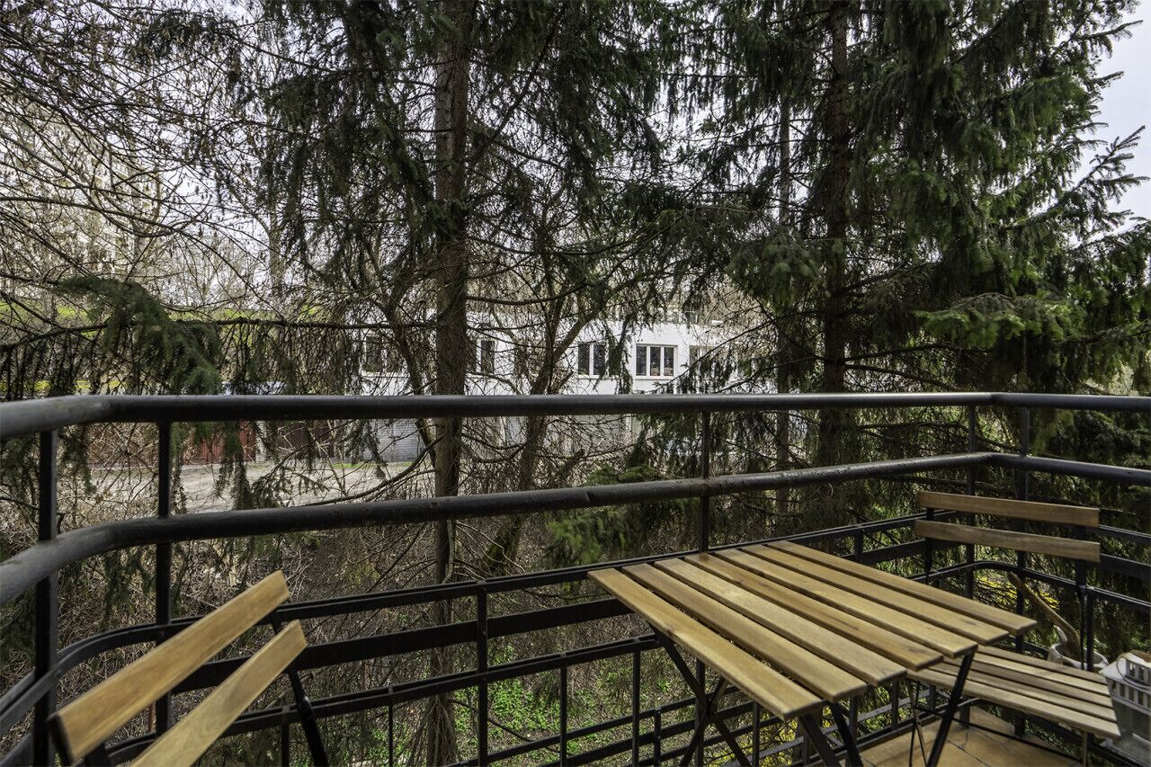A wooden bench on a balcony with a metal railing, overlooking a forested area and a white building in the background.