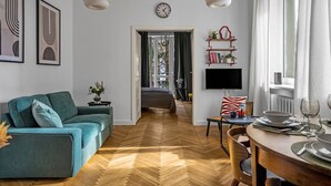 Un salon moderne avec un canapé, un coin repas avec une table dressée pour deux personnes, un téléviseur fixé au mur et un couloir menant à une chambre.