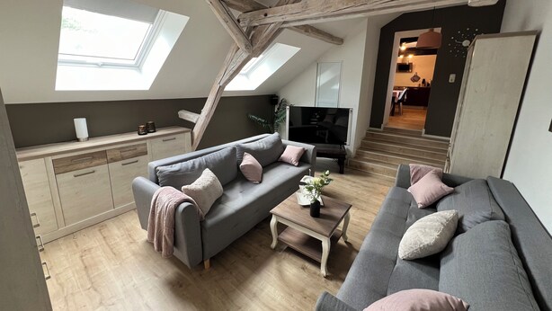En stue i loftstil med højt til loftet, synlige bjælker, ovenlys, sofaarrangement, sofabord og et fjernsyn.