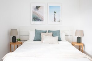 Cama bem arrumada com lençóis brancos, duas almofadas e uma pequena planta em vaso sobre uma mesa de cabeceira.