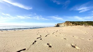 Ein sandiger Strand mit Fußspuren, die zum Meer führen, und eine Klippe im Hintergrund.