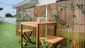 Una mesa de madera y dos sillas en un patio con césped artificial, una valla de madera con plantas trepadoras y un edificio blanco al fondo.