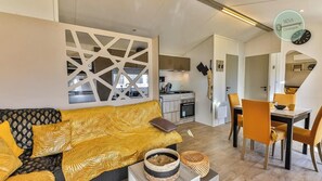 A modern living room with a sofa, a dining table, and a kitchen in the background. - Mobil-home dans parc résidentiel (Saint-Hilaire-de-Riez)