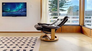 A modern room with a leather recliner, a large window with a view of trees, and a painting of the Northern Lights on the wall. - Waterfront villa near IKEA and Sylvia Park (Auckland)