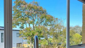 A kitchen with a stainless steel sink and a window overlooking a backyard with trees and a wooden fence. - Waterfront villa near IKEA and Sylvia Park (Auckland)