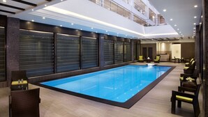 A modern indoor swimming pool with a glass ceiling, lounge chairs, and a reception area. - The Radiant Swan (Vagra)