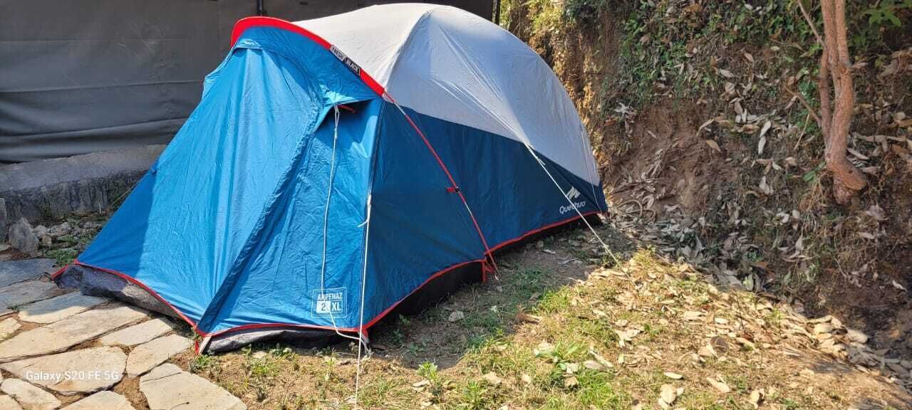 A blue and gray tent with a red trim, set up on a stone path beside a dirt area with sparse vegetation.