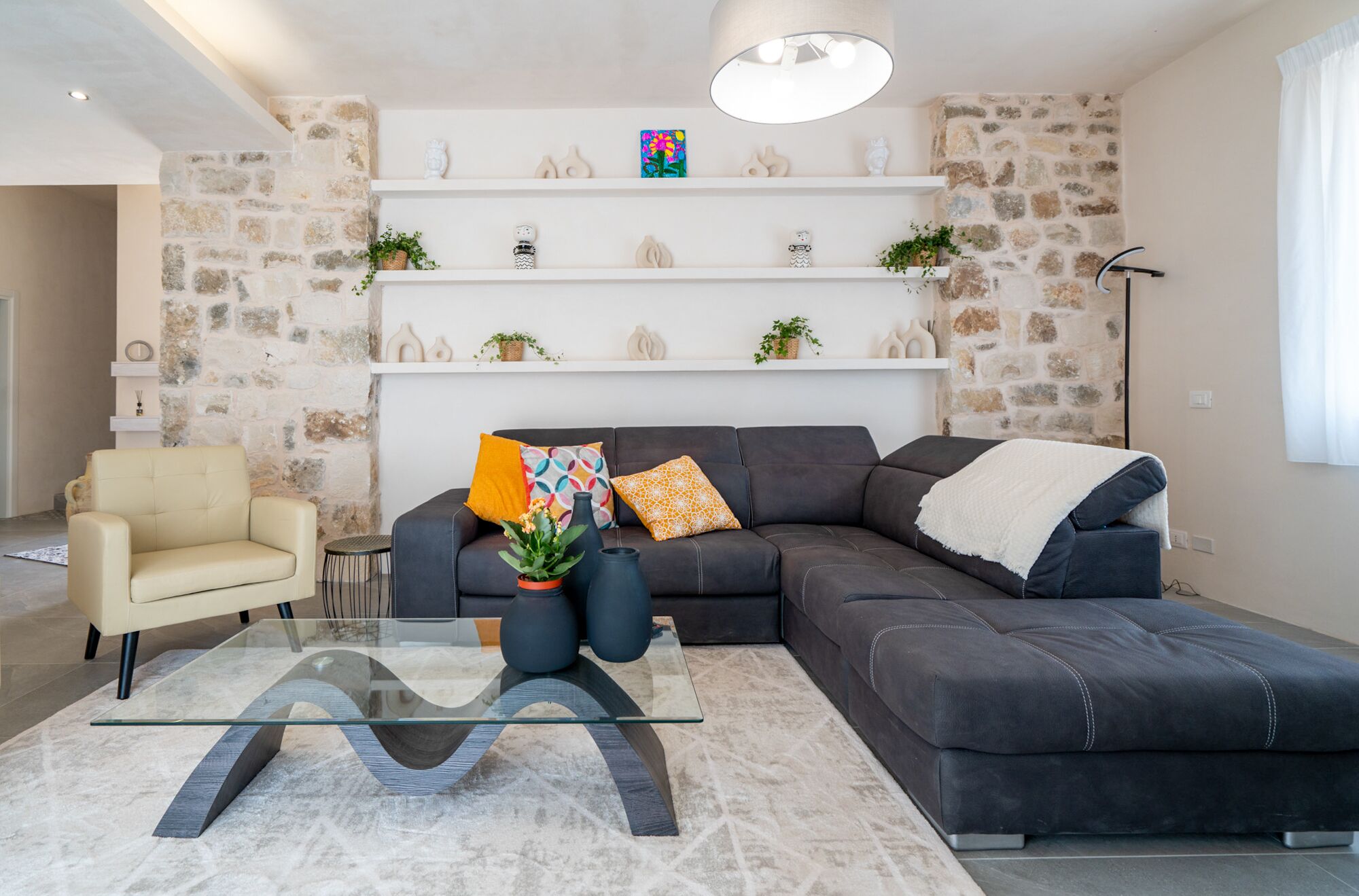 A modern living room with a sectional sofa, a glass coffee table, and decorative items on shelves against a stone wall.