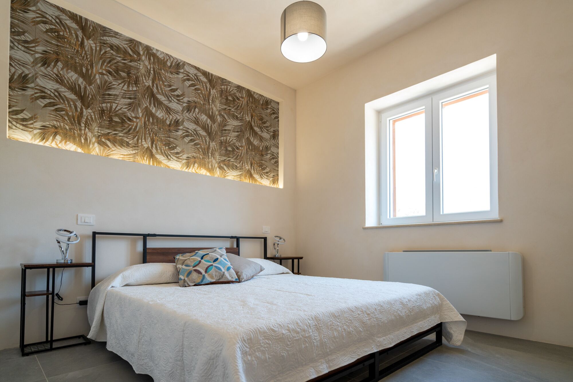 A bedroom with a bed, a nightstand, a lamp, a radiator, and a window with blinds.