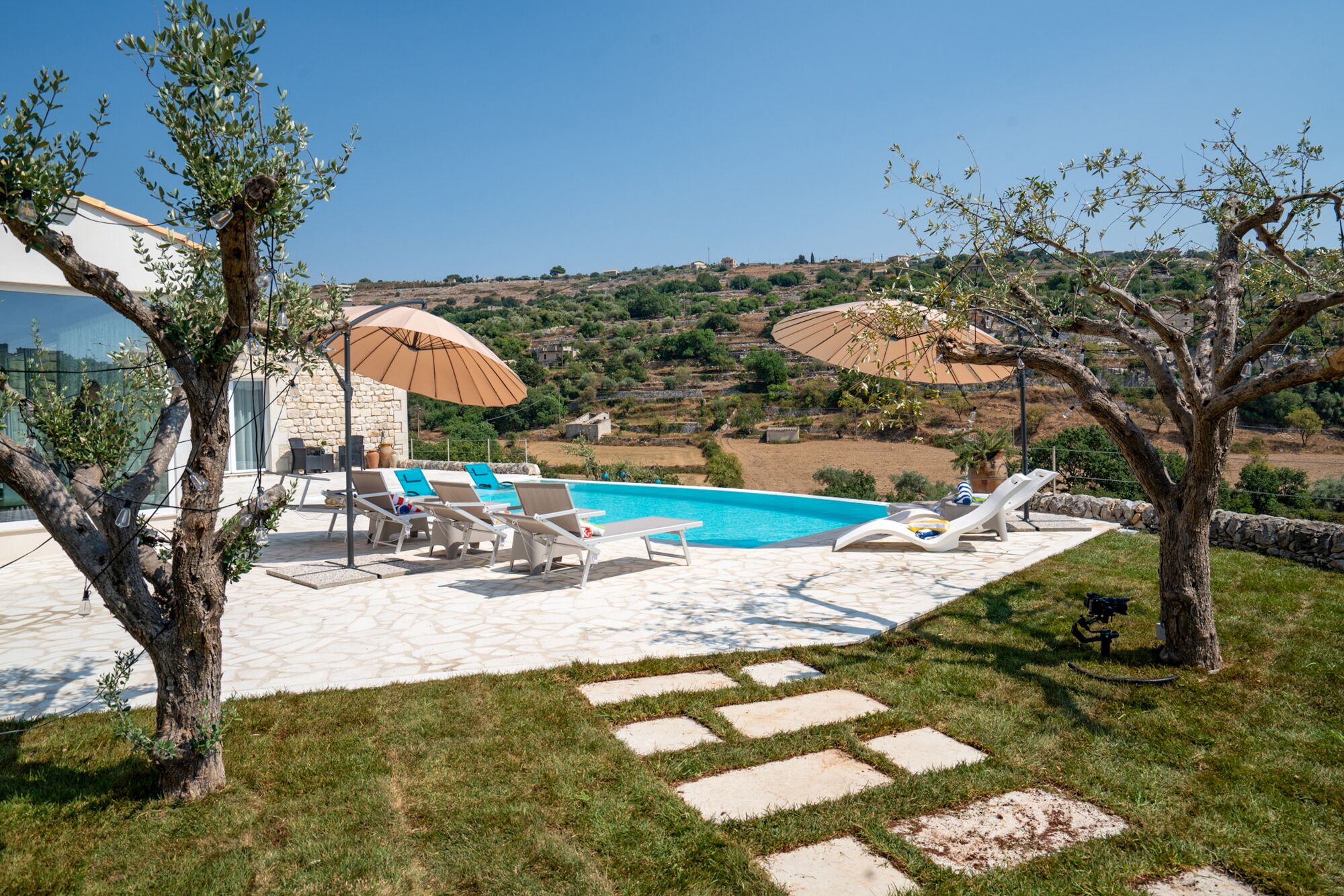 A pool area with lounge chairs and umbrellas, surrounded by a stone pathway and grass.