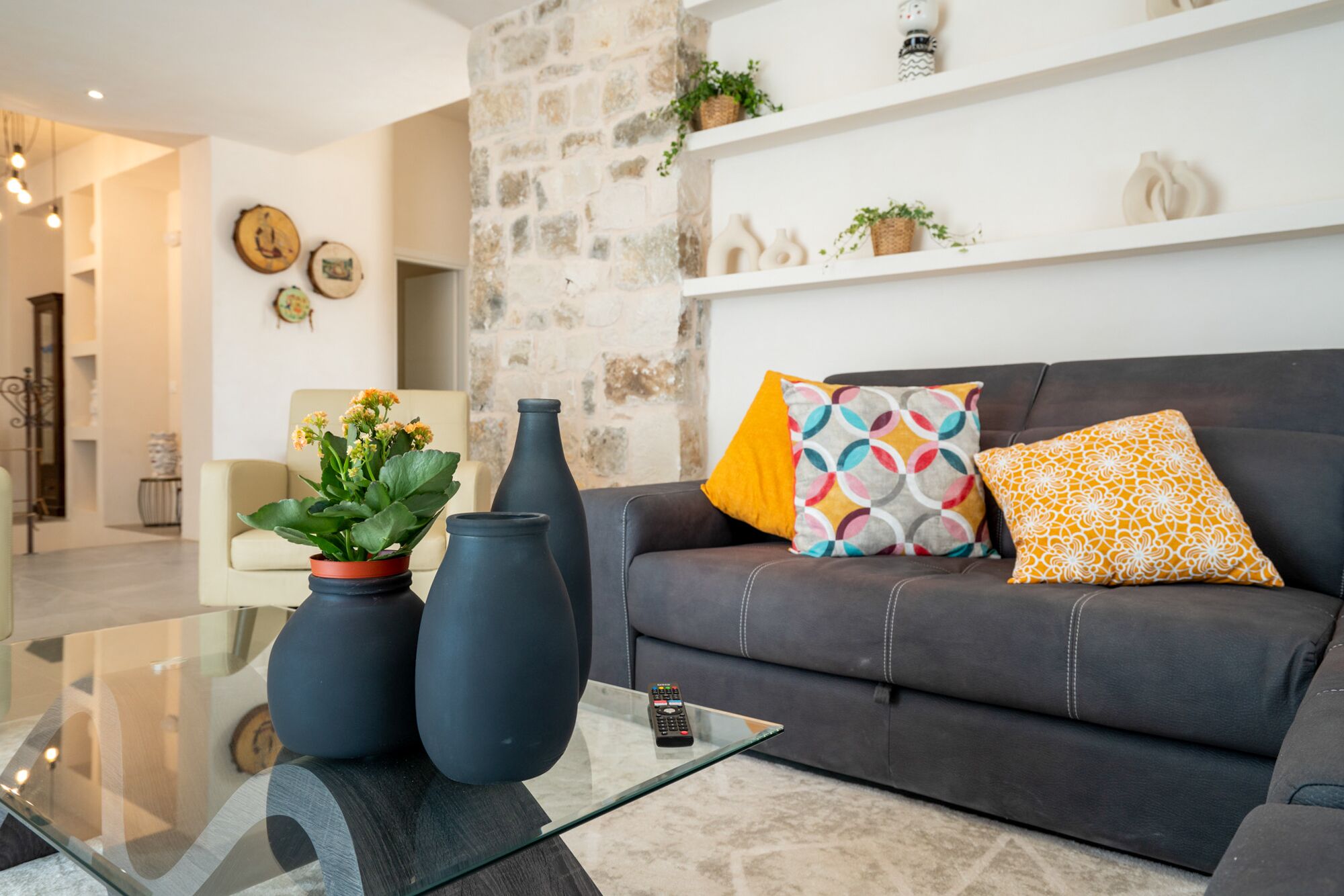 A modern living room with a dark leather sofa, colorful cushions, a glass coffee table, and decorative vases.