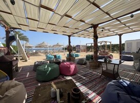 Et udendørs opholdsområde med farverige pufsofaer og en stribet måtte, med udsigt over en strand med parasoller og en klar himmel.